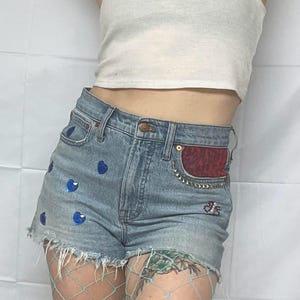 May include: Light blue denim shorts with a frayed hem and blue heart embellishments. The shorts have a red and black leopard print patch on the back pocket.