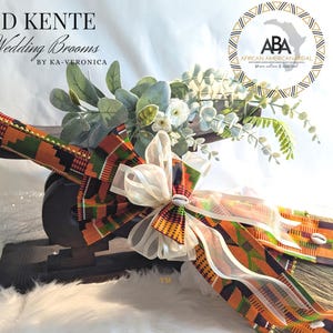 Cultured Kente Wedding Broom By African American Bridal is shown with traditional vibrant orange, black and white Kente cloth with gold beading and cowrie shells on the broom spray laying on a traditional African Stool for display.
