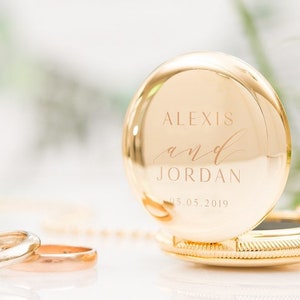 May include: A gold pocket watch with the names "Alexis and Jordan" and the date "05.05.2019" engraved on the cover. Two gold wedding bands are in the foreground. The watch is open, revealing the inner workings.