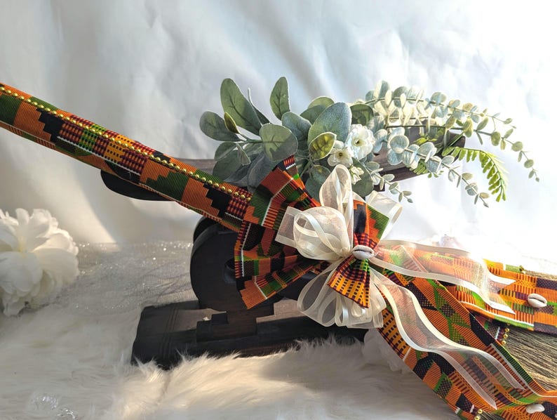 Close-up of a cultured Kente wedding broom by African American Bridal showcasing traditional Kente fabric, symbolic cowrie shells, gold bead accents, and a sheer ivory bow used for Jumping the Broom ceremonies