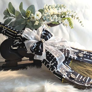 Handcrafted cultured Kente wedding broom by African American Bridal, featuring vibrant black and white Kente cloth, cowrie shell accents, gold bead detailing, and an white organza bow, designed to honor the African American wedding tradition of jumping the broom. 🧹