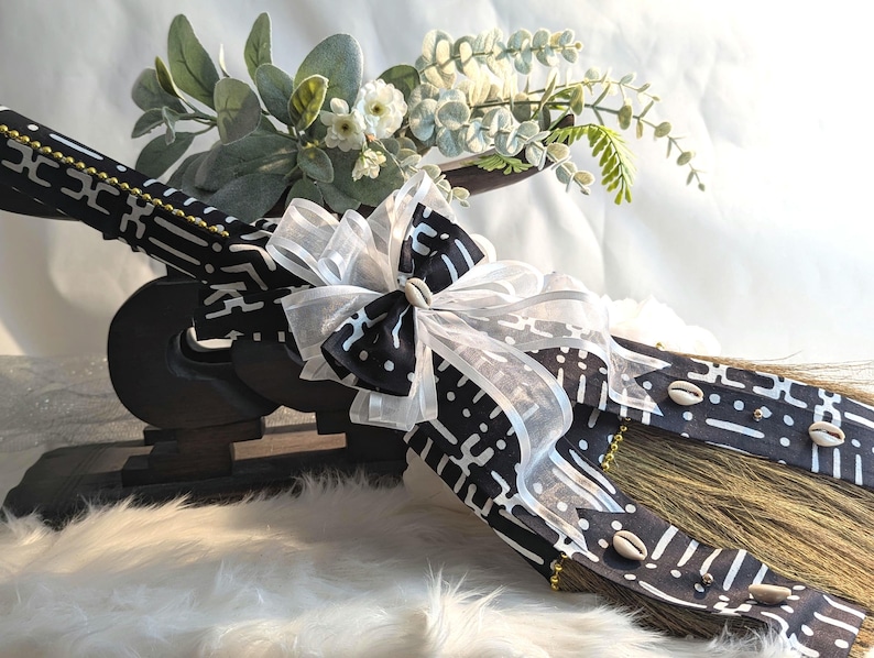 Handcrafted cultured Kente wedding broom by African American Bridal, featuring vibrant black and white Kente cloth, cowrie shell accents, gold bead detailing, and an white organza bow, designed to honor the African American wedding tradition of jumping the broom. 🧹