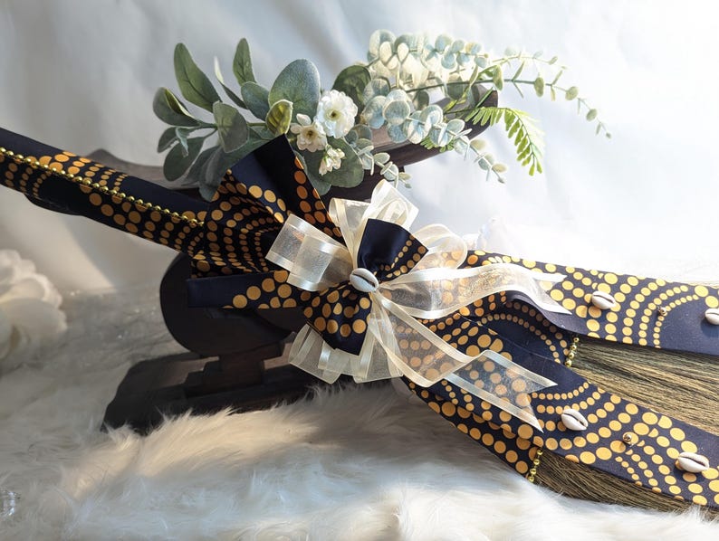 Handcrafted cultured Kente wedding broom by African American Bridal, featuring vibrant multicolor Kente cloth, cowrie shell accents, gold bead detailing, and an ivory organza bow, designed to honor the African American wedding tradition of jumping the broom. Ethnic Fabics available Black & Gold cultured bling Wedding Broom 🧹