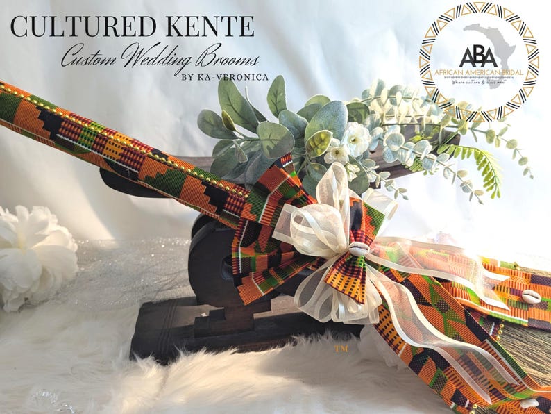 Cultured Kente Wedding Broom By African American Bridal is shown with traditional vibrant orange, black and white Kente cloth with gold beading and cowrie shells on the broom spray laying on a traditional African Stool for display.