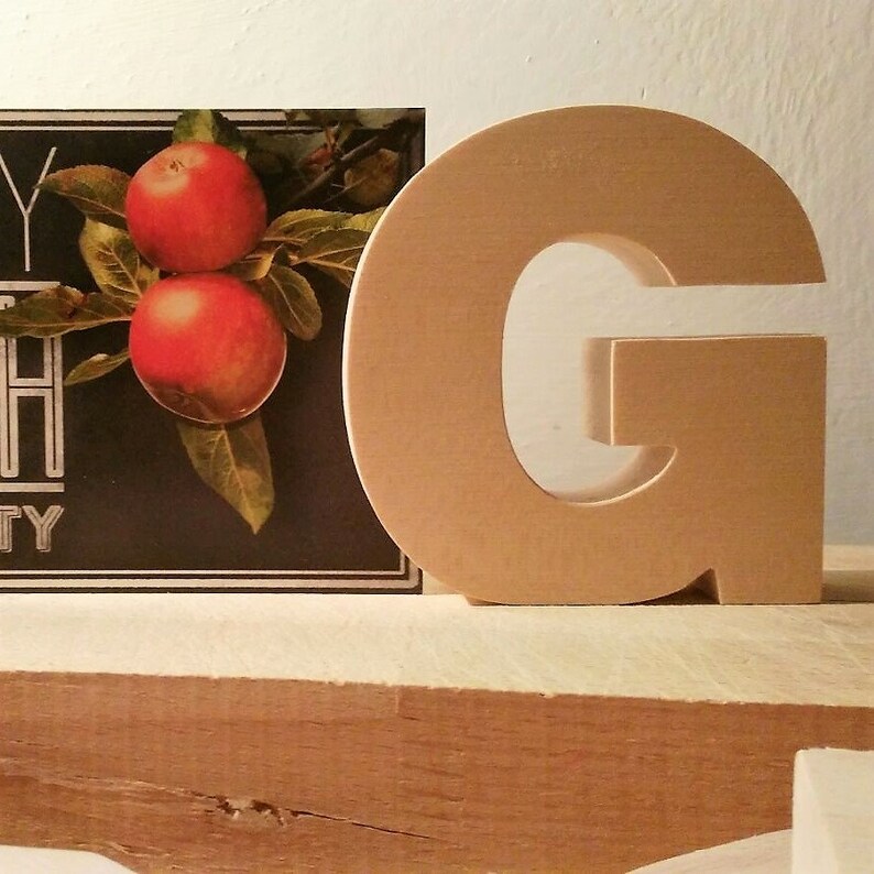 May include: A wooden letter G, painted in a light brown color, sits on a wooden surface. The letter is freestanding and has a simple, classic design.