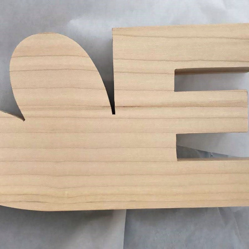 May include: Unpainted wooden letters spelling "LOVE" with a heart shape for the "O". The letters are made of natural wood with a smooth finish.