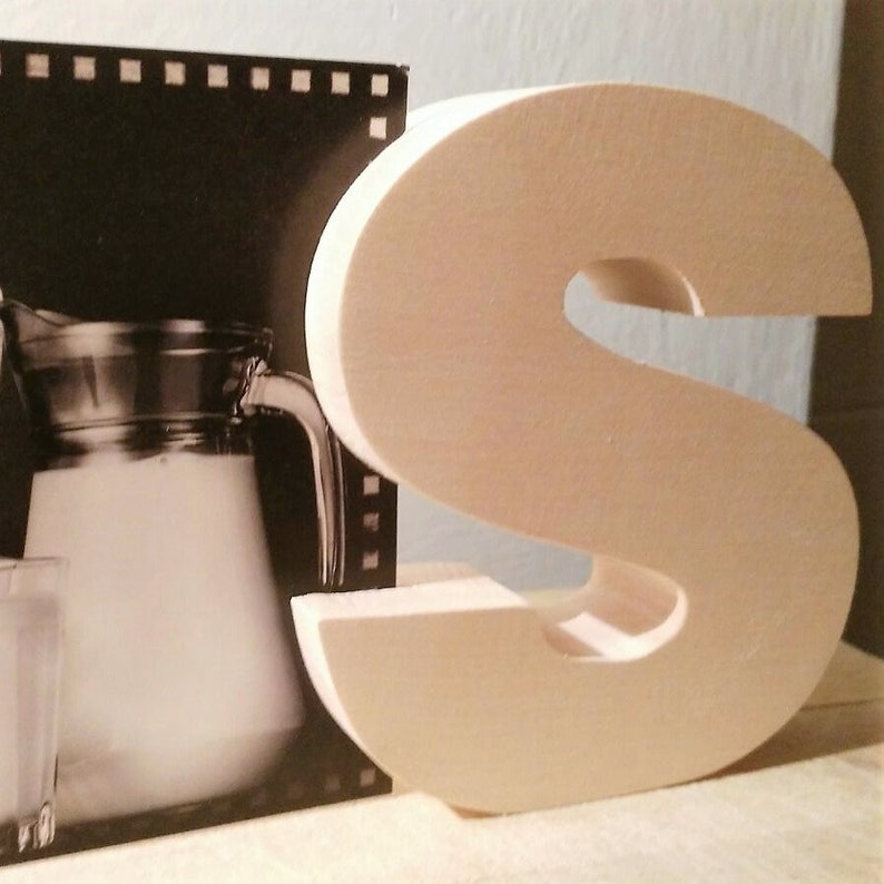 May include: A large, wooden, light brown letter S. The letter is standing on a surface with a black and white photo of a pitcher of milk in the background.