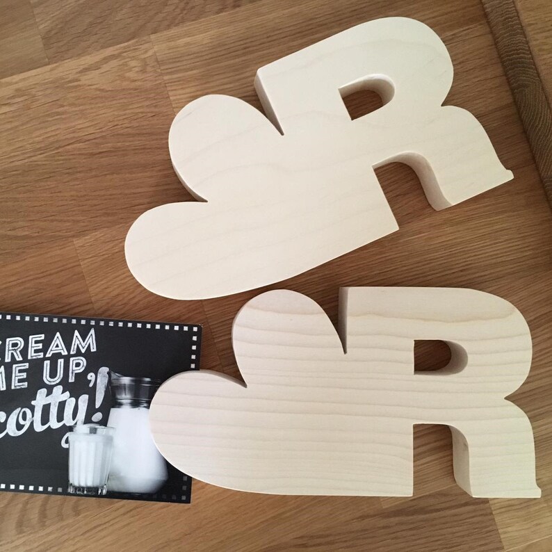 May include: Two unfinished wooden letters, both are the letter R. The letters are in a heart shape. The letters are on a wooden surface.