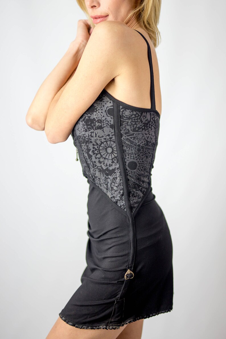 May include: Black dress with a decorative gray panel featuring a sun and moon pattern. The dress has thin straps and a fitted bodice that flows into a skirt. A gold-colored clasp accents the side.