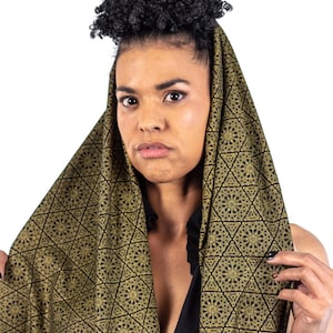 May include: A woman with curly hair is wearing a black and gold patterned scarf. The scarf is draped over her head and shoulders, and the pattern is intricate and geometric.