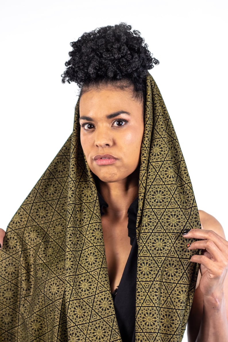 May include: A woman with curly hair is wearing a black and gold patterned scarf. The scarf is draped over her head and shoulders, and the pattern is intricate and geometric.