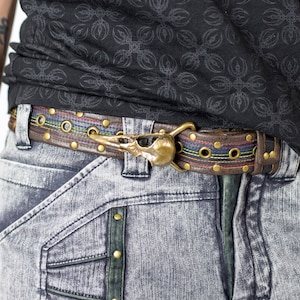 Może przedstawiać: Brązowy skórzany pasek z mosiężną klamrą w kształcie czaszki ptaka. Pasek ma kolorowe przeszycia i mosiężne oczka. Noszony z szarymi dżinsami.