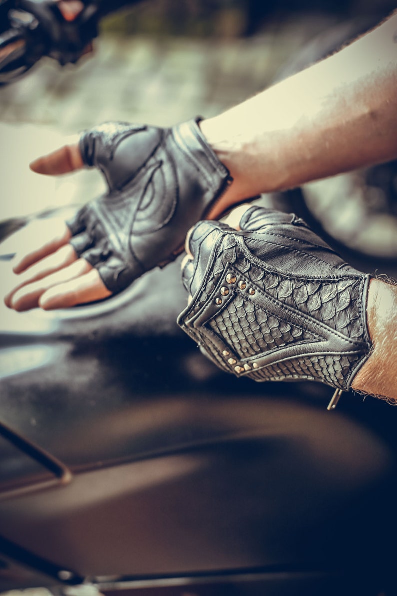K&ouml;nnte beinhalten: Ein Paar schwarze Leder-Motorradhandschuhe mit fingerlosem Design. Ein Handschuh hat ein strukturiertes Muster und silberne Nieten.