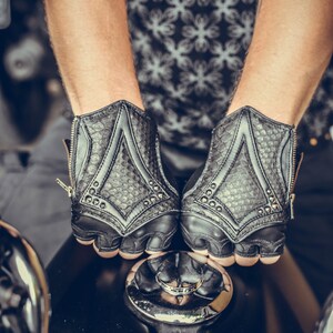 K&ouml;nnte beinhalten: Ein Paar schwarze Lederfingerlose Handschuhe mit einem strukturierten Muster und silbernen Nieten. Die Handschuhe haben einen Rei&szlig;verschluss auf der R&uuml;ckseite des Handgelenks.