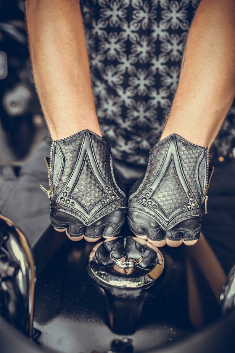 K&ouml;nnte beinhalten: Ein Paar schwarze Lederfingerlose Handschuhe mit einem strukturierten Muster und silbernen Nieten. Die Handschuhe haben einen Rei&szlig;verschluss auf der R&uuml;ckseite des Handgelenks.