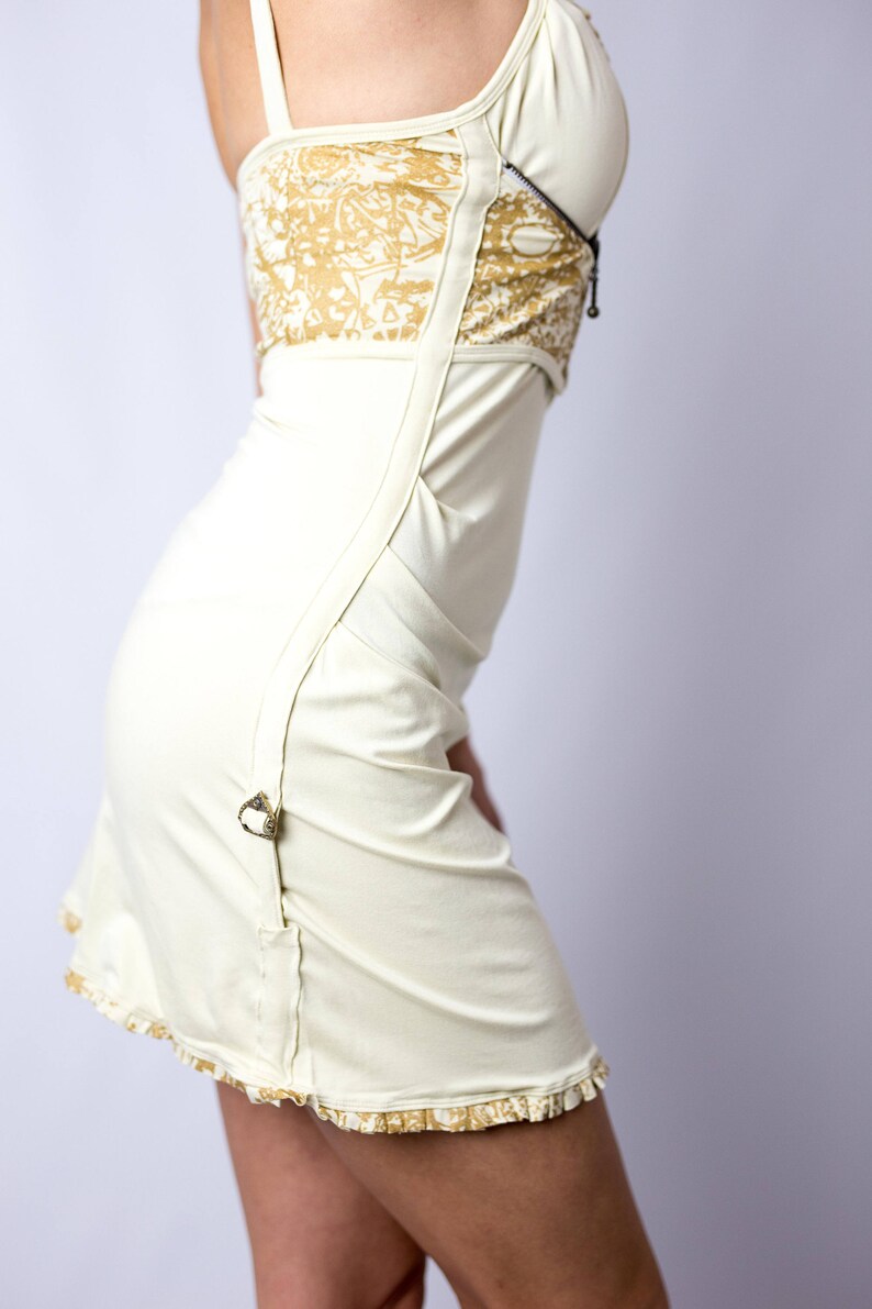 May include: Ivory-colored dress with a fitted bodice and a flared skirt. The bodice features a gold patterned panel and a zipper detail. The dress has thin straps and decorative buckles. The hemline is trimmed with a gold patterned ruffle.