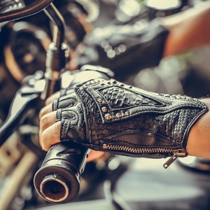 K&ouml;nnte beinhalten: Nahaufnahme eines schwarzen Ledermotorradhandschuhs mit silbernen Nieten und einem Rei&szlig;verschluss. Der Handschuh wird von einer Person getragen, die ein Motorrad f&auml;hrt.