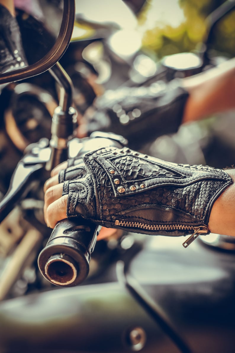 K&ouml;nnte beinhalten: Nahaufnahme eines schwarzen Ledermotorradhandschuhs mit silbernen Nieten und einem Rei&szlig;verschluss. Der Handschuh wird von einer Person getragen, die ein Motorrad f&auml;hrt.