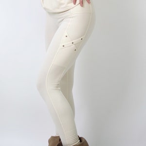 May include: White leggings with a decorative detail on the left thigh. The detail is a criss-cross pattern with small metal studs. The leggings are worn with brown ankle boots.