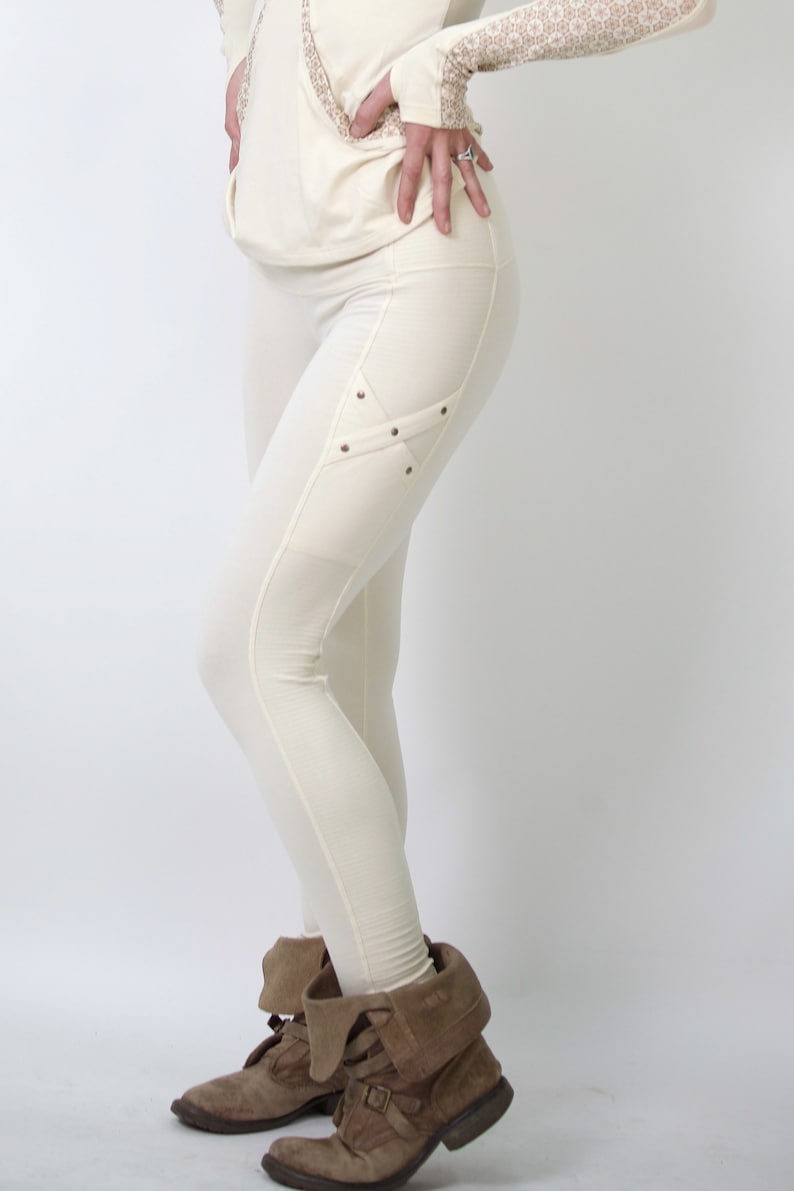 May include: White leggings with a decorative detail on the left thigh. The detail is a criss-cross pattern with small metal studs. The leggings are worn with brown ankle boots.