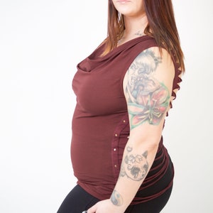 May include: A woman wearing a maroon cowl neck top with a side lace-up detail and black pants. She has a large tattoo on her right arm.