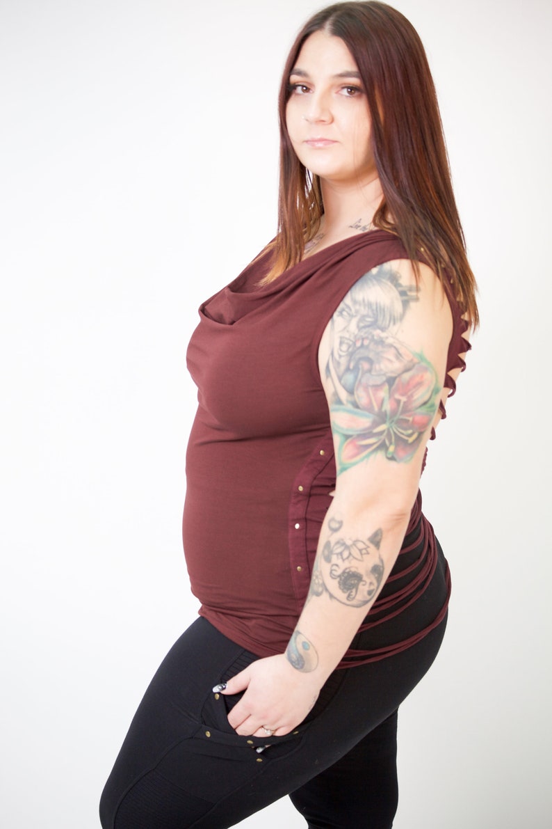 May include: A woman wearing a maroon cowl neck top with a side lace-up detail and black pants. She has a large tattoo on her right arm.
