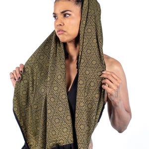May include: A woman wearing a black and gold patterned scarf with a geometric design. The scarf is draped over her head and shoulders, and she is looking to the side.