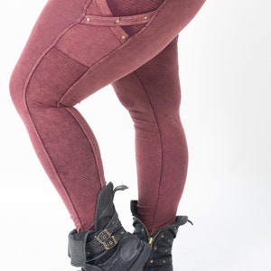 May include: Burgundy coloured leggings with a decorative pocket on the left thigh. The pocket has a criss-cross design with gold accents. The leggings are worn with black leather boots.