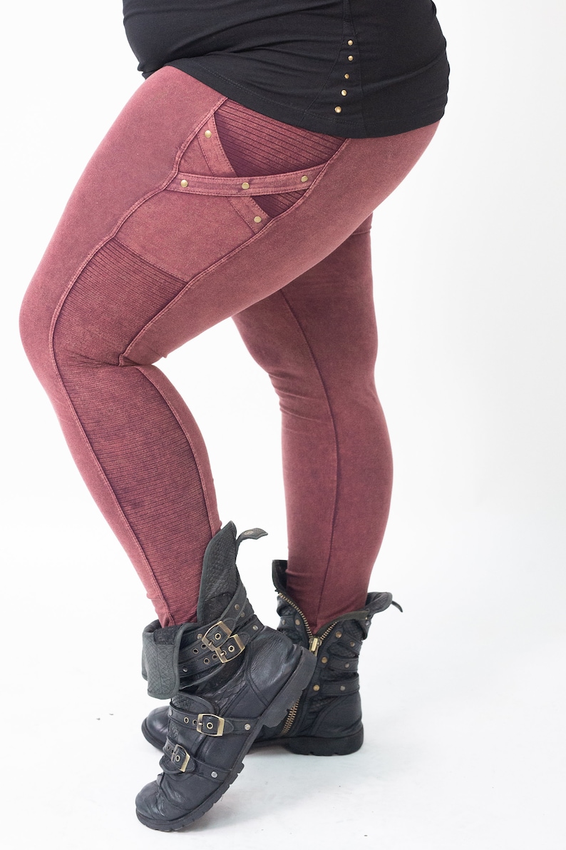 May include: Burgundy coloured leggings with a decorative pocket on the left thigh. The pocket has a criss-cross design with gold accents. The leggings are worn with black leather boots.