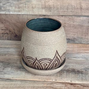 May include: A round ceramic planter with a speckled beige exterior and a dark teal interior. The lower portion of the planter features a dark brown etched pattern of repeating petal shapes. It sits on a matching circular ceramic saucer.