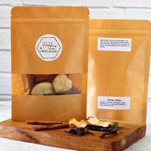 May include: Two brown paper pouches with a clear window, filled with dried fruit and herbs, sit on a wooden board. Cinnamon sticks and other ingredients are scattered around. One pouch has a label that says "Winter Glow". The other pouch has a sticker that says "Holoka Home".