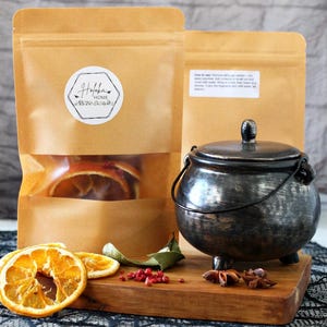 May include: A still-life image featuring a small, black cauldron with a lid, set on a wooden board with dried orange slices, spices, and two brown paper bags. One bag has a clear window showing orange slices and a label.