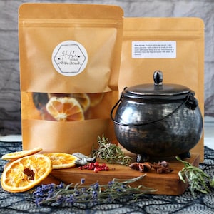 May include: A still-life image featuring a small, dark metal cauldron with a lid, next to two brown paper pouches. One pouch has a clear window showing dried orange slices. The scene includes a wooden board with spices and herbs.
