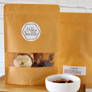 May include: A brown paper bag with a white label that says "Holoka Home" and a clear window showing dried apple slices, pink peppercorns, lavender, and cloves. The bag is labeled "Salvatore" and contains the text "Contains: dried apples, pink peppercorns, lavender, cloves, star anise (NOT FOR CONSUMPTION)".