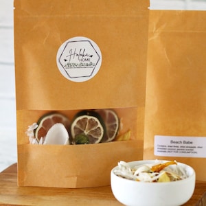 May include: A brown paper bag with a white label that says "Haloka Home" and a clear window showing dried fruit and lime slices. The bag is sitting on a wooden surface next to a white bowl filled with dried fruit and coconut flakes. The text "Beach Babe" is visible on the second bag.