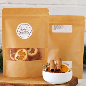 May include: Two brown paper pouches with a clear window showing dried orange slices and cinnamon sticks. A small white bowl holds more dried oranges, cinnamon sticks, and rosemary. The logo on the pouch says "Hofka Home".
