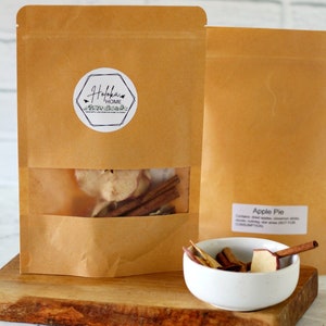 May include: A brown paper bag with a clear window showing dried apple slices, cinnamon sticks, and other spices. The bag has a white sticker with the text "Hohoha Home" on it. The bag is sitting on a wooden surface next to a white bowl filled with dried apple slices and cinnamon sticks. The bowl is labeled "Apple Pie".