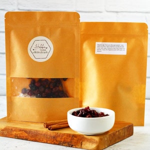 May include: Two brown paper pouches with a clear window showing dried berries. A small white bowl filled with berries and two cinnamon sticks sit on a wooden cutting board. The pouches have a sticker that says "Hoffka Home".