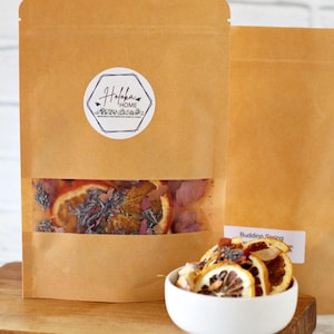 May include: Brown paper bag with a clear window displaying dried orange slices, lavender, and rose petals. A white bowl holds a similar mix. The bag has a white label with the text "Holoka Home". A wooden cutting board is in the foreground.