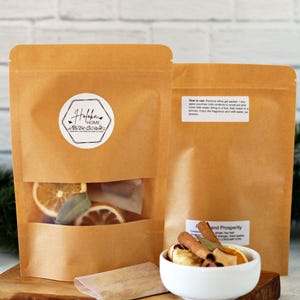 May include: Two brown paper pouches with a clear window showing dried orange slices, bay leaves, and cinnamon sticks. A small white bowl contains similar ingredients. The label on the pouch reads "Hofoka Home".