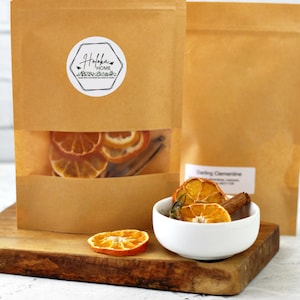 May include: Two brown paper bags with a white label that says "Haloka Home" and dried orange slices and cinnamon sticks inside. A white bowl with dried orange slices and cinnamon sticks is on a wooden cutting board.