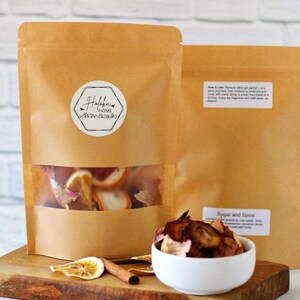 May include: Two brown paper bags with a white label that says "Haloka Home" and a clear window showing dried fruit inside. The bags are on a wooden surface with a white bowl of dried fruit and a cinnamon stick.