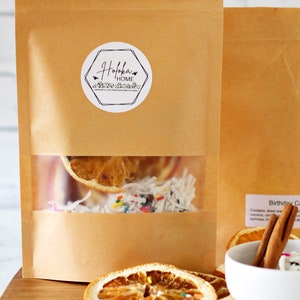 May include: Kraft paper pouches with a clear window displaying dried orange slices, coconut flakes, and colorful sprinkles. The pouches have a sticker with the text "Holoka Home". A small white bowl holds cinnamon sticks and more ingredients.