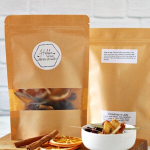 May include: Two brown paper pouches with clear windows, filled with dried fruit and spices. One pouch has a label that says "Hofka's Home". A small white bowl contains a drink with fruit and spices. Cinnamon sticks and orange slices are on a wooden board.
