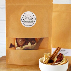 May include: Brown paper bag with a clear window, filled with dried fruit and a cinnamon stick. A small white bowl contains a similar mix. The bag has a circular label with the text "Holoka Home".
