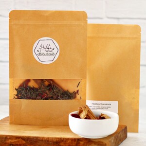 May include: Two brown paper pouches of tea, one with a clear window showing the tea blend. A white bowl holds cinnamon sticks and other tea ingredients. The tea is labeled "Holiday Romance" with ingredients listed.