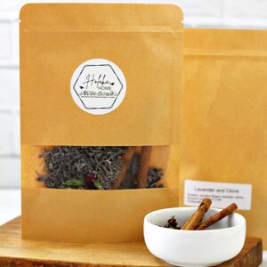 May include: A brown paper bag with a white sticker that says "Holoka Home" and a clear window showing dried lavender, cinnamon sticks, and other herbs. A white bowl with cinnamon sticks and dried flowers sits on a wooden cutting board.