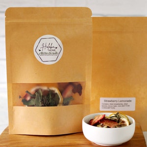 May include: A brown paper bag with a clear window showing dried fruit and herbs inside. The bag has a white sticker with the text "Holokai Home" on it. A white bowl with dried fruit and herbs is on a wooden surface. The text "Strawberry Lemonade" is printed on the bag.