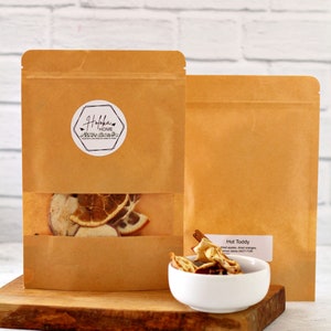 May include: Two brown paper bags, one with a clear window showing dried orange slices and other ingredients. A small white bowl contains dried fruit and cinnamon sticks. The label on the bag reads "Hot Toddy".