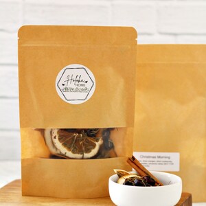 May include: Brown paper bags with a clear window, filled with dried citrus slices, rosemary, and berries. A small white bowl contains similar ingredients, with a cinnamon stick. The bags have a sticker that says "Hofoka Home".
